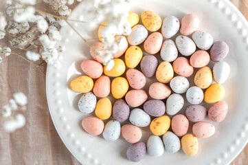 Colorful Easter chocolate eggs in a plate with flowers on a beige background, top view.