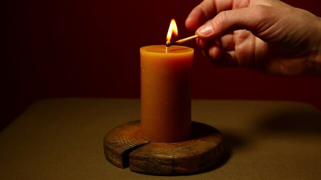 Woman hand lights a match the bees wax candle on wooden candlestick for mood, relaxation, meditation dark red background. Person lighting the decoration single candle flame