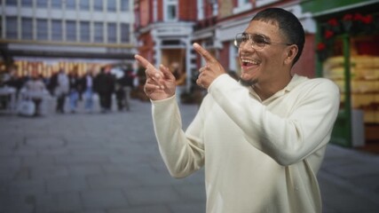 Man wearing glasses points both index fingers to the left while smiling on a crowded city street with storefronts and holiday stalls; playful joy.