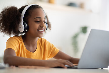 Cheerful black girl studying online, using laptop and headset at home, typing on keyboard, copy space. Cute african american teen kid having online lesson, kitchen interior. Home schooling concept