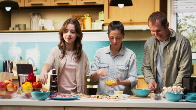 Pre-teen daughter taking pictures of her and parents preparing pizza together, making memories through fun weekend activity. Girl takes a family photo in the kitchen, bonding. Camera B.