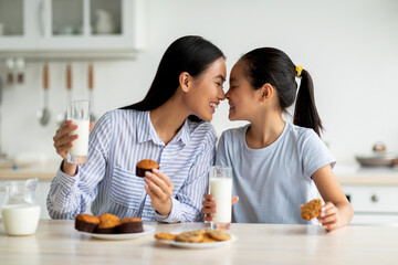 Loving asian mother and little daughter having snacks and drinking milk in kitchen interior,...