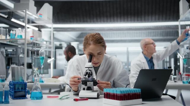 Female microscopist examining microscope glass slide in laboratory to view cells. Research facility employee checks sample slide, doing imaging process using optical device, camera A