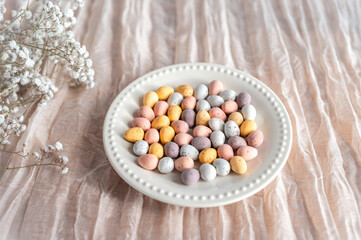 Colorful Easter chocolate eggs in a plate with flowers on a beige background, top view.