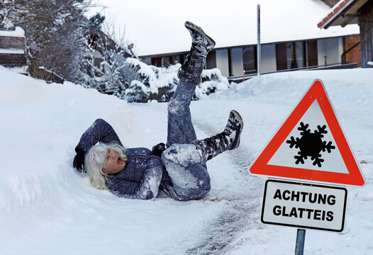 Rutschgefahr auf einem glatten Gehweg im Winter. Eine Frau ist auf dem Eis ausgerutscht. Schild mit Achtung Glatteis 