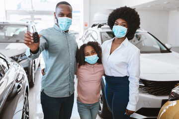 Portrait of black family in face masks showing car key, buying new vehicle in auto dealership during covid lockdown. Afro parents with cute kid purchasing automobile at showroom