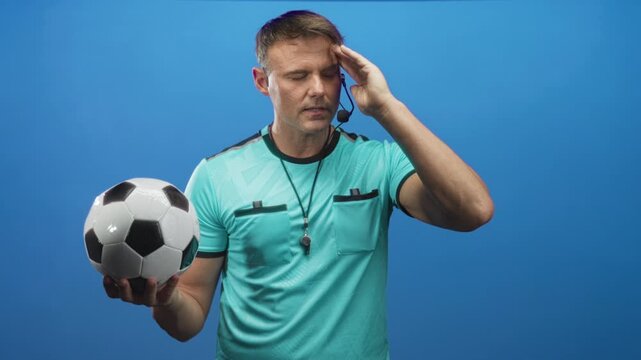 Man holding a soccer ball in one hand and touching his temple with the other hand, referee with whistle and headset visible in studio; focus strategy.
