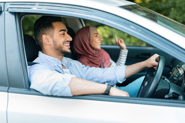 Glad smiling millennial islamic husband driving at steering wheel with wife in hijab, ride in car, enjoy music while traveling. Fun in road trip, vacation, love, relationship and family vacation
