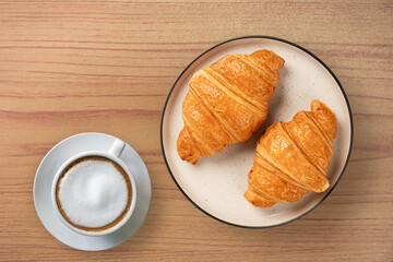 Top- view of two golden-brown crispy croissants served on a ceramic plate alongside a creamy cup of cappuccino. A perfect breakfast set on a warm wooden background.