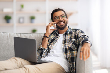 Joyful middle-eastern young man freelancer working from home, sitting on couch with laptop, having...