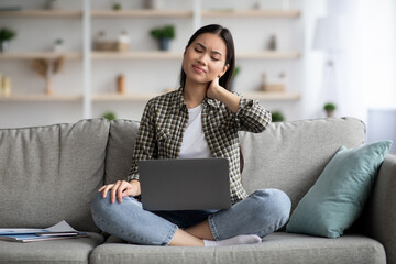 Upset asian young woman in casual suffering from neck pain while sitting on couch and using laptop,...