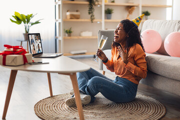 Obraz premium Distance Birthday Party. Joyful Black Lady At Laptop Celebrating B-Day Online Drinking Champagne Having Group Video Call With Friends Sitting At Home, Wearing Festive Hat. Side View Shot