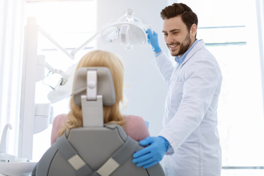 Young handsome bearded man dentist doctor greeting and comforting female patient sitting in dentist chair, turning on lamp adn smiling, wearing rubber gloves, copy space, modern dental clinic interior