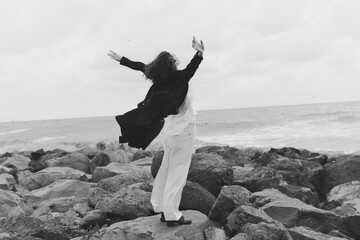 Freedom, celebration, ocean on rocky coastline with couple raising arms in motion, wind sweeps garments, black and white scene capturing spontaneous joy and adventurous spirit