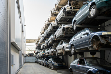 Stacked cars in scrapyard with multiple vehicles lined up on shelves. The scene shows rows of cars awaiting recycling or dismantling on sunny day 