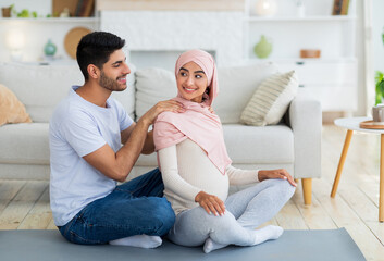 Loving arab husband massaging his pregnant wife, woman feeling relaxed, spouses exercising together on mat, sitting on floor at living room, copy space