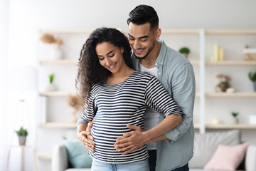 Fototapeta premium Loving middle eastern young man husband hugging his beautiful pregnant wife big tummy, millennial arabic family waiting for baby, home interior, copy space. Family, childbirth, pregnancy concept