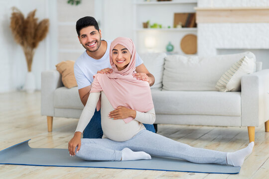 Pregnancy workout. Happy arab spouses exercising together at home, young expectant woman stretching legs while man massaging wife's shoulders and smiling together to camera - Powered by Adobe