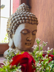 Peaceful buddha statue head indoors with natural light and flowers creating calm meditation atmosphere and spiritual decorative home scene. 