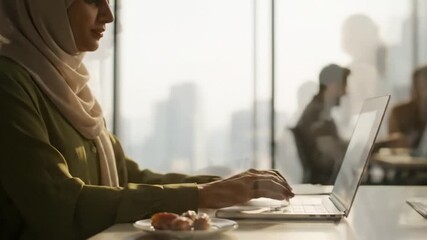 Candid video of Muslim professional in modern office preparing online meeting during Ramadan, placing dates, drinking water, typing laptop, calm productivity mood, subtle camera movement.
