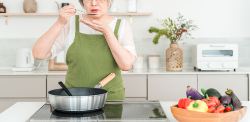 A senior woman in an apron tasting food while cooking in her home kitchen