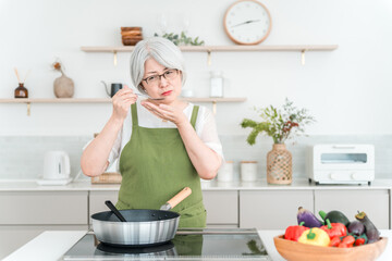 A senior woman in an apron tasting food while cooking in her home kitchen