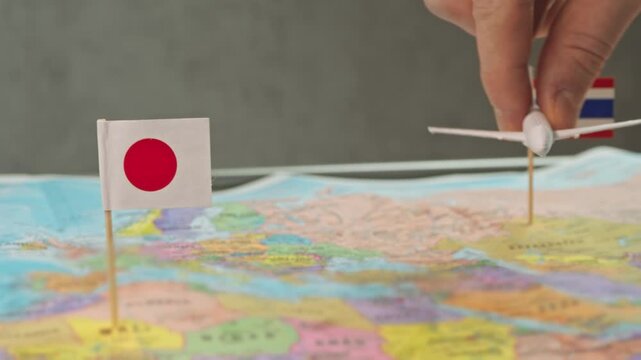 Toy airplane flying from Thailand to Japan above colorful world map, symbol cherry blossom tours, winter vacations, cultural exchange trips and popular seasonal air travel between countries.