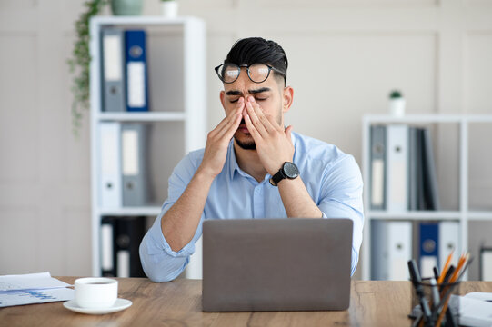 Millennial Arab employee rubbing tired irritated eyes, working with laptop too much, exhausted from online job at office. Young Eastern businessman feeling fatigue from looking at screen