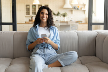 Happy young woman sitting on couch in living room and using cellphone, checking new mobile application at home, copy space. Millennial lady surfing web, having video chat with friend on smartphone