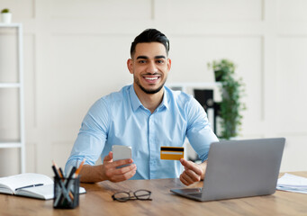 Online shopping, web store concept. Cheerful young Arab man holding credit card and cellphone, making remote payment, ordering goods on web, buying on internet from modern office
