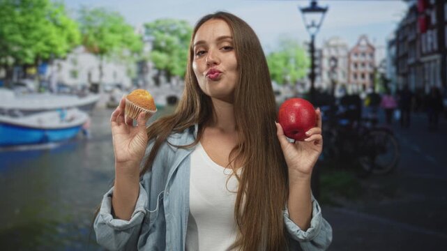 Woman holding apple and muffin on street in amsterdam by a canal and lamppost, wearing denim jacket and smiling; playful choice.