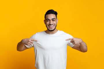 Excited arab guy pointing with both hands at his blank white t-shirt and smiling, posing over...