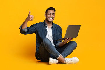 Cheerful bearded young arabic guy in casual outfit sitting on floor with modern laptop and showing...