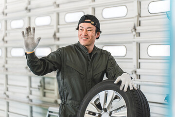 A middle-aged Asian man working as a car mechanic greeting someone while holding a tire
