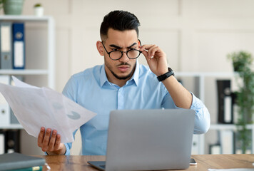 Obraz premium Stressed young Arab employee using laptop, holding documents, having problem with work task, making mistake in business project at office. Millennial guy having trouble meeting deadline