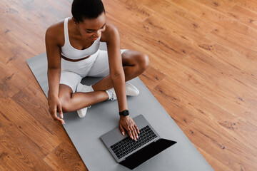 Sport And Recreation Concept. Above top high angle view of smiling fit black woman watching fitness videos on internet using laptop typing on keyboard sitting on fitness mat in living room