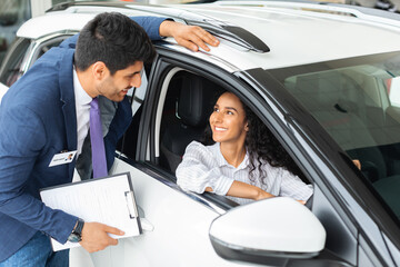 Pretty long-haired brunette lady sitting inside brand new comfy white car, having conversation with...