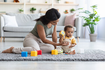Obraz premium Young black mother and her baby boy playing with toys at home, sitting on floor in living room interior, happy african american mom and infant child enjoying spending time together, copy space