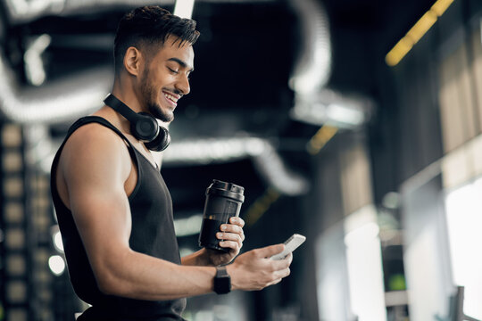 Fitness App. Handsome Smiling Arab Sporty Man Using Smartphone While Standing In Gym, Happy Middle Eastern Male Athlete Browsing New Mobile Application For Workout Productivity Tracking, Copy Space