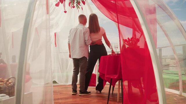 Couple enjoying rooftop igloo date, candlelit table, red curtains, panoramic city view at sunset, intimate embrace and champagne toast, cozy dome atmosphere, whispered conversation and romantic mood.