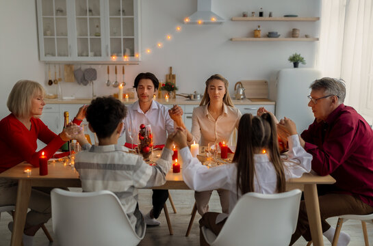 Multi generation family holding hands and praying before Thanksgiving or Christmas dinner, sitting at table indoors. Kids, their parents and grandparents worshiping God, thankful for festive meal