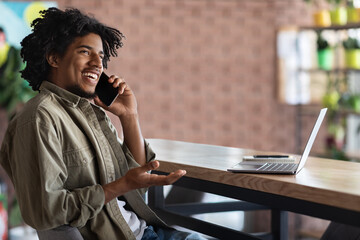 Smiling millennial african american curly man talking by smartphone sitting at table with laptop, notepad in cafe interior. Work remote for independent contractor, call to client at coworking space