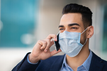 Closeup portrait of young arab businessman in protective medical face mask talking on cellphone next to business center, copy space. Muslim entrepreneur or CEO working during COVID-19 pandemic