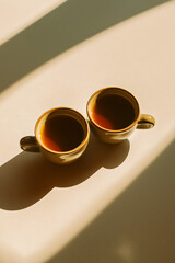Two golden coffee cups on a beige surface with sunlight