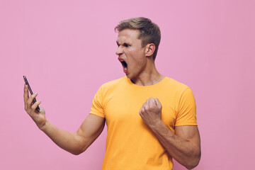 Full body portrait of a young angry man with light skin, short hair, wearing a yellow t-shirt, expressing frustration while looking at a smartphone, isolated on plain pink background, studio shot