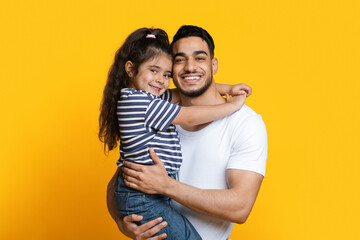 Naklejka premium Portrait Of Happy Young Arab Dad Holding His Little Daughter On Hands, Loving Middle Eastern Dad Bonding With Cute Female Child While They Posing Together Over Yellow Studio Background, Copy Space