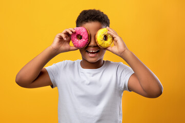 Funny afro american preteen boy in white t-shirt holding colorful delicious donuts over eyes, cheerful black guy having fun while enjoying tasty sweets over yellow studio background