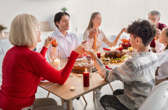Multi generation family praying before holiday dinner, holding hands, giving thanks to God at home. Young couple with kids and senior parents celebrating Thanksgiving or Christmas - Powered by Adobe