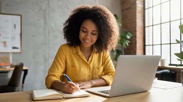 Young woman working on her laptop and writing in a notebook in a stylish workspace