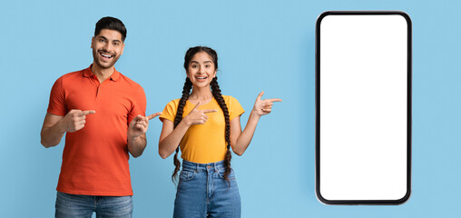 A young man and woman are happily posing with gestures, pointing at a blank smartphone screen. They are dressed casually against a bright blue background, showcasing positivity.